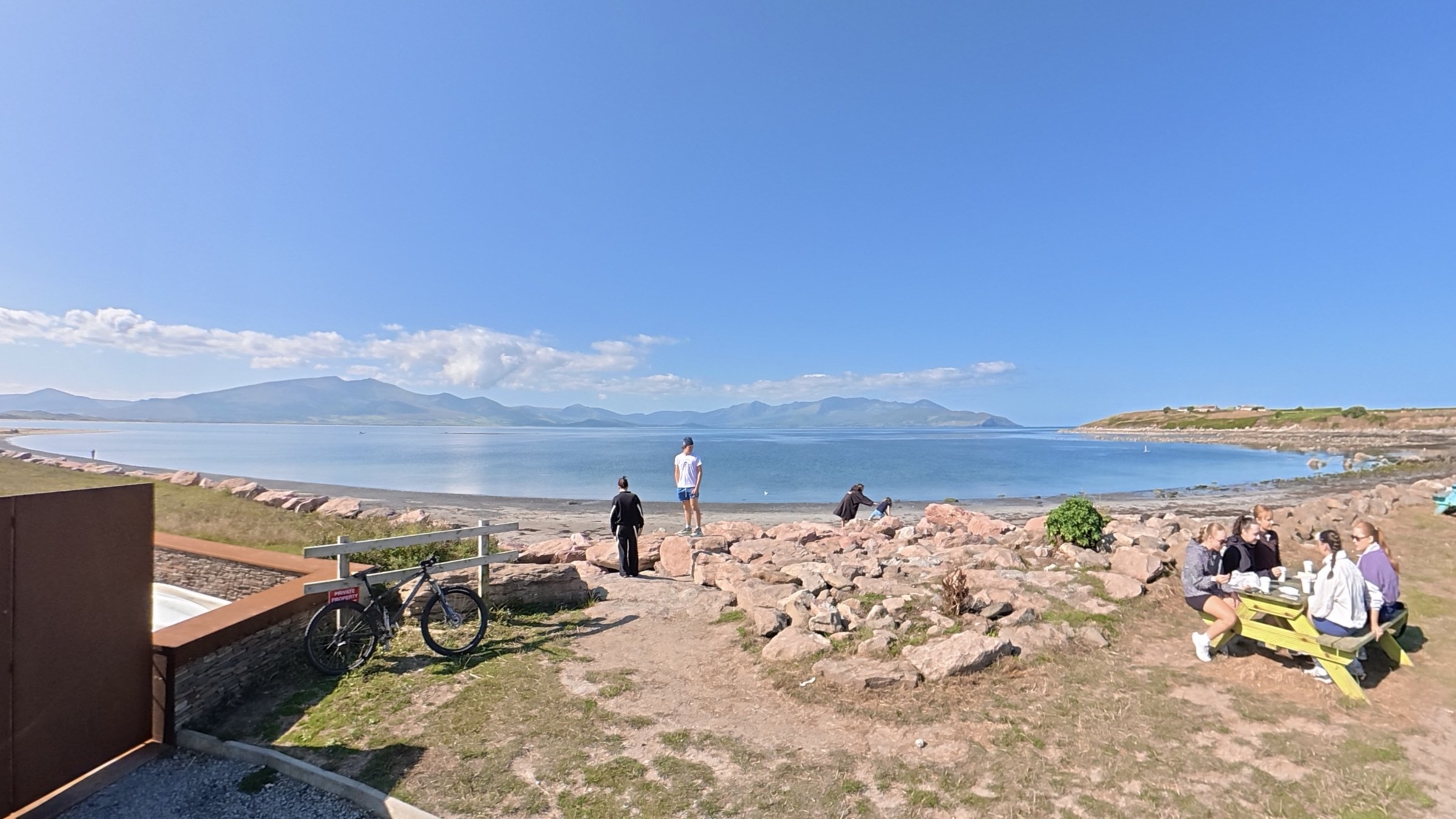 People sitting among rocks beside the calm blue water at the Maharees